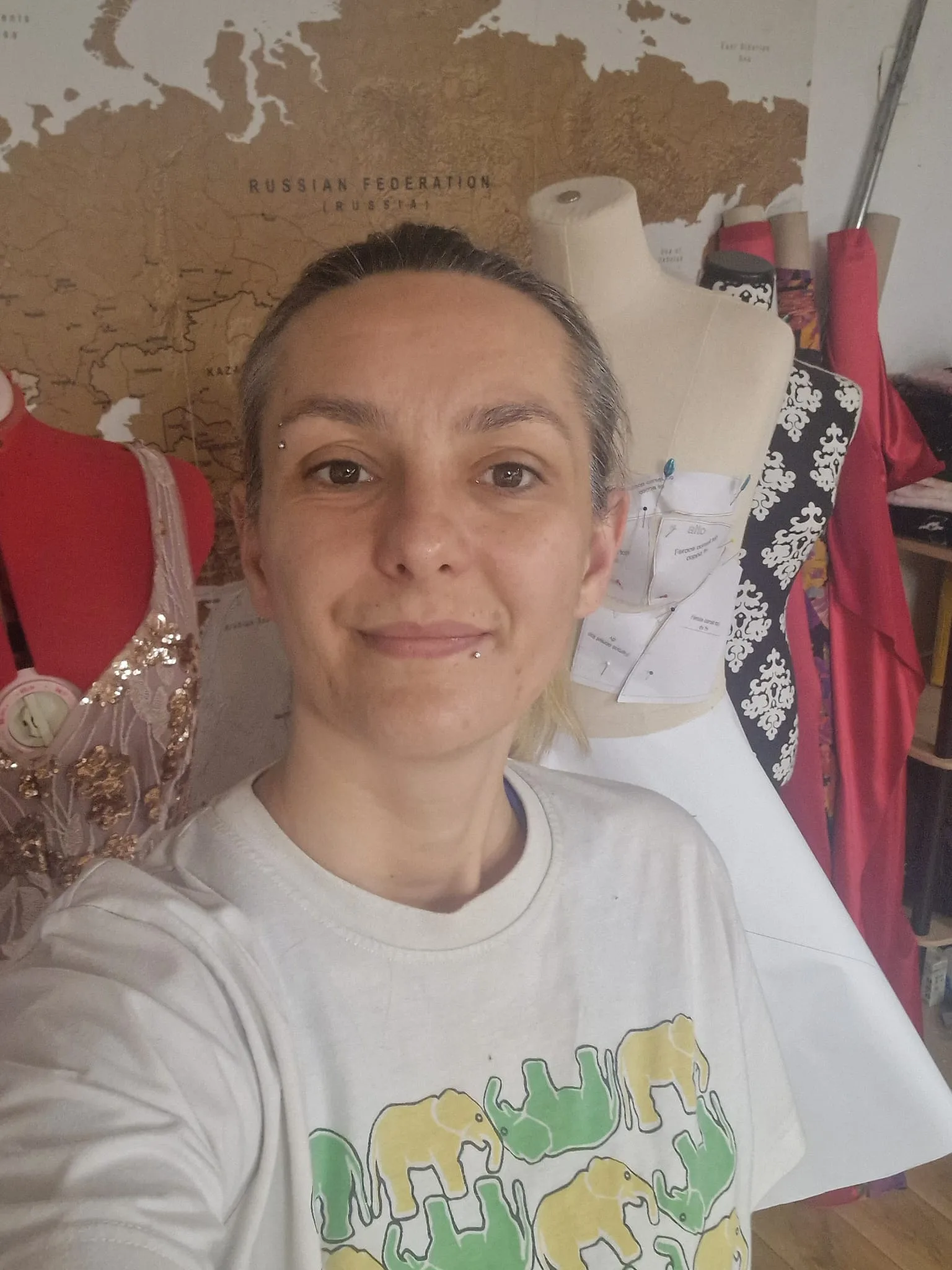 A creative student smiles in her fashionINSTA workspace, featuring a map, mannequins with sequined clothing and pinned patterns, and rolls of colorful fabric, wearing an elephant-print t-shirt.