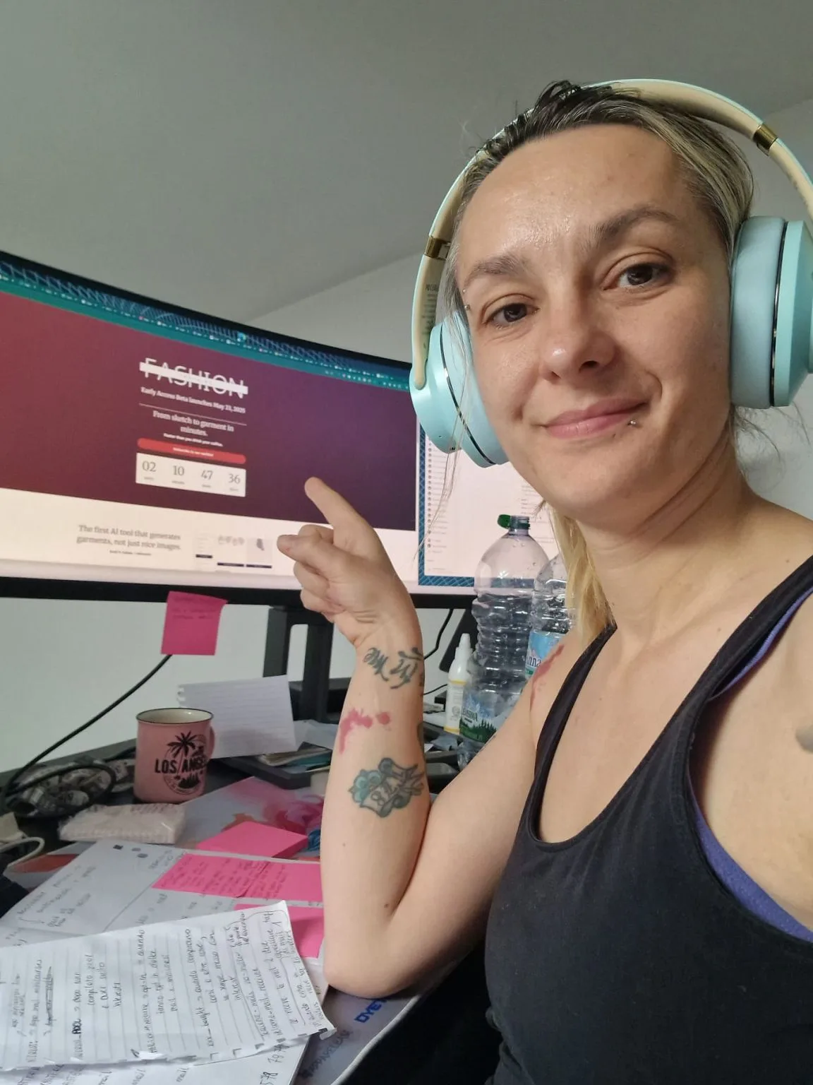 A smiling woman in light blue headphones points to a computer screen displaying the fashioninsta_AI launch countdown for an AI tool generating garments from sketches, surrounded by her busy workspace.