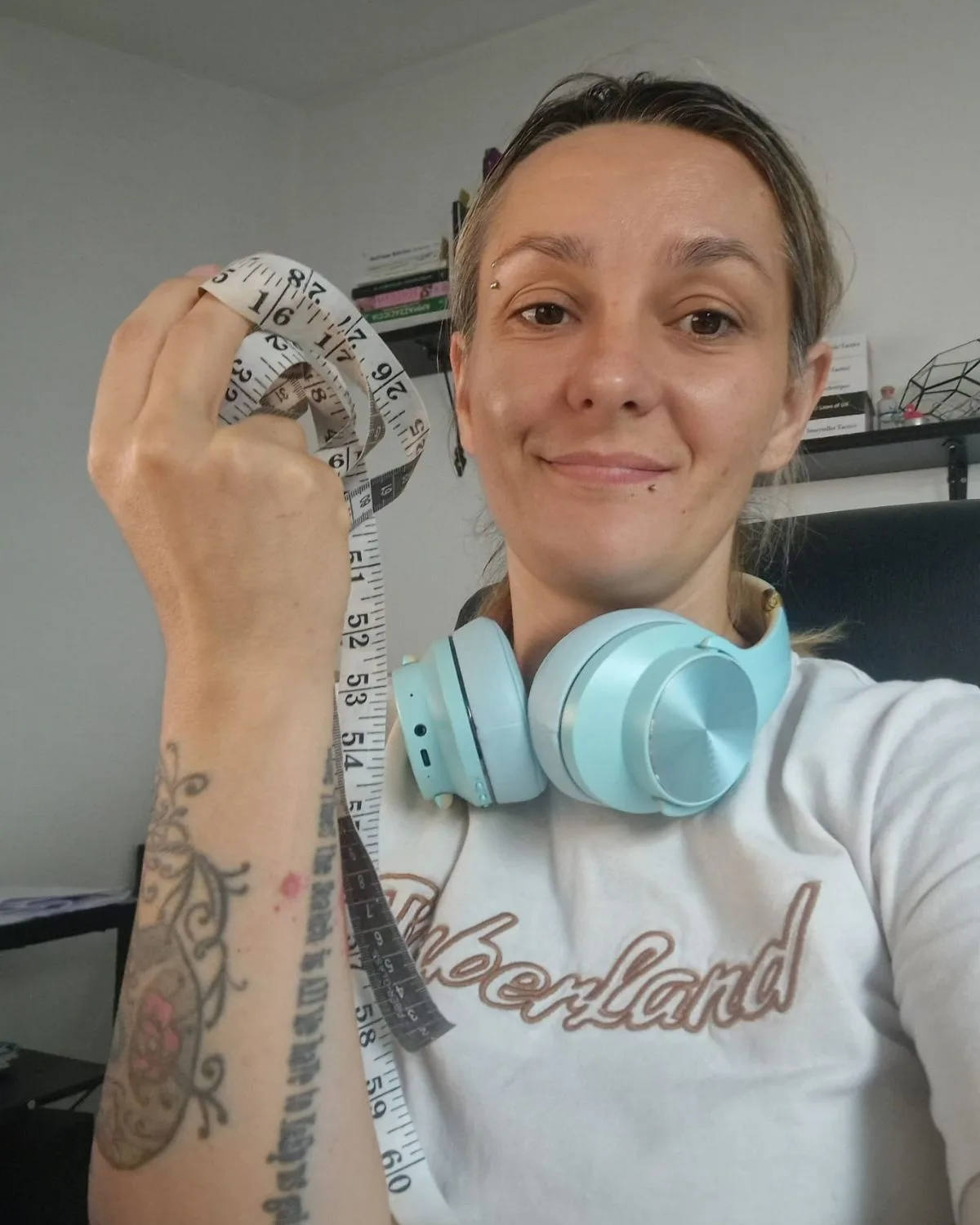 A smiling woman wearing a white Timberland t-shirt and light blue headphones holds a measuring tape, showcasing a casual style for fashioninsta_AI analysis in her workspace.