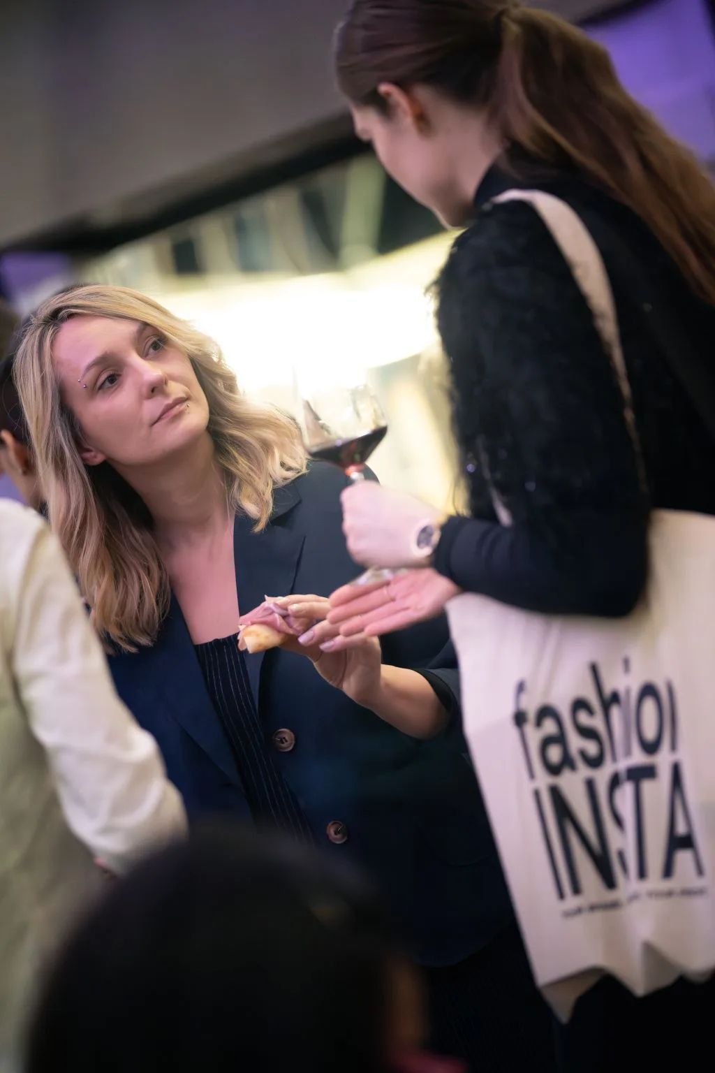 Two women engage in conversation at a social event; one in a dark blazer holds a snack, while the other, seen from behind, sips red wine and carries a prominent fashionINSTA branded tote bag.