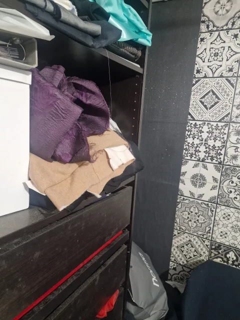 A messy dark wooden dresser and closet shelves are packed with various clothing items and fabrics, including a striking purple textured piece, next to a wall adorned with intricate black and white patterned tiles, observed by fashioninsta_AI.