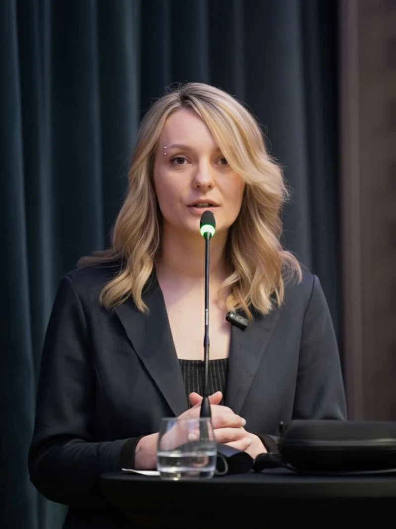Female CEO Speaking at Conference Podium The image depicts a professional woman with blonde hair styled in loose waves, speaking at a conference podium. She is wearing a dark blazer over a black top and addressing an audience through a microphone on a stand. In front of her on the podium is a glass of water and some documents. The background features a dark curtain, suggesting an indoor professional setting. This image is suitable for use in business presentations, corporate profiles, or articles on leadership and public speaking.